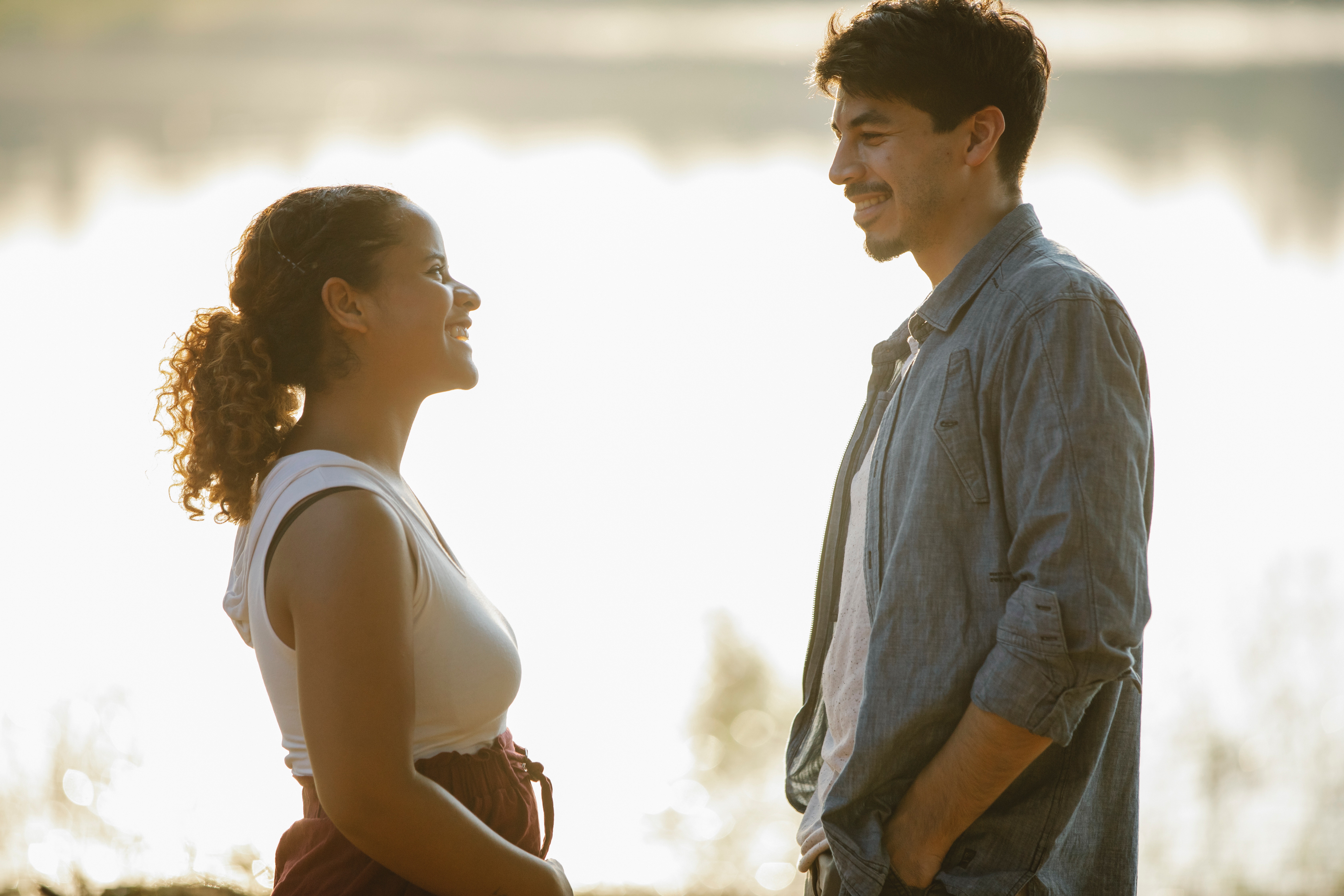 An attractive man and woman talk while smiling. They are relaxed and standing in front of a lake.