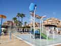 Ein Kinder-Planschbecken mit Wasserrutsche und Springbrunnen, umgeben von Sonnenliegen und Sonnenschirmen vor einer Resort-Kulisse.