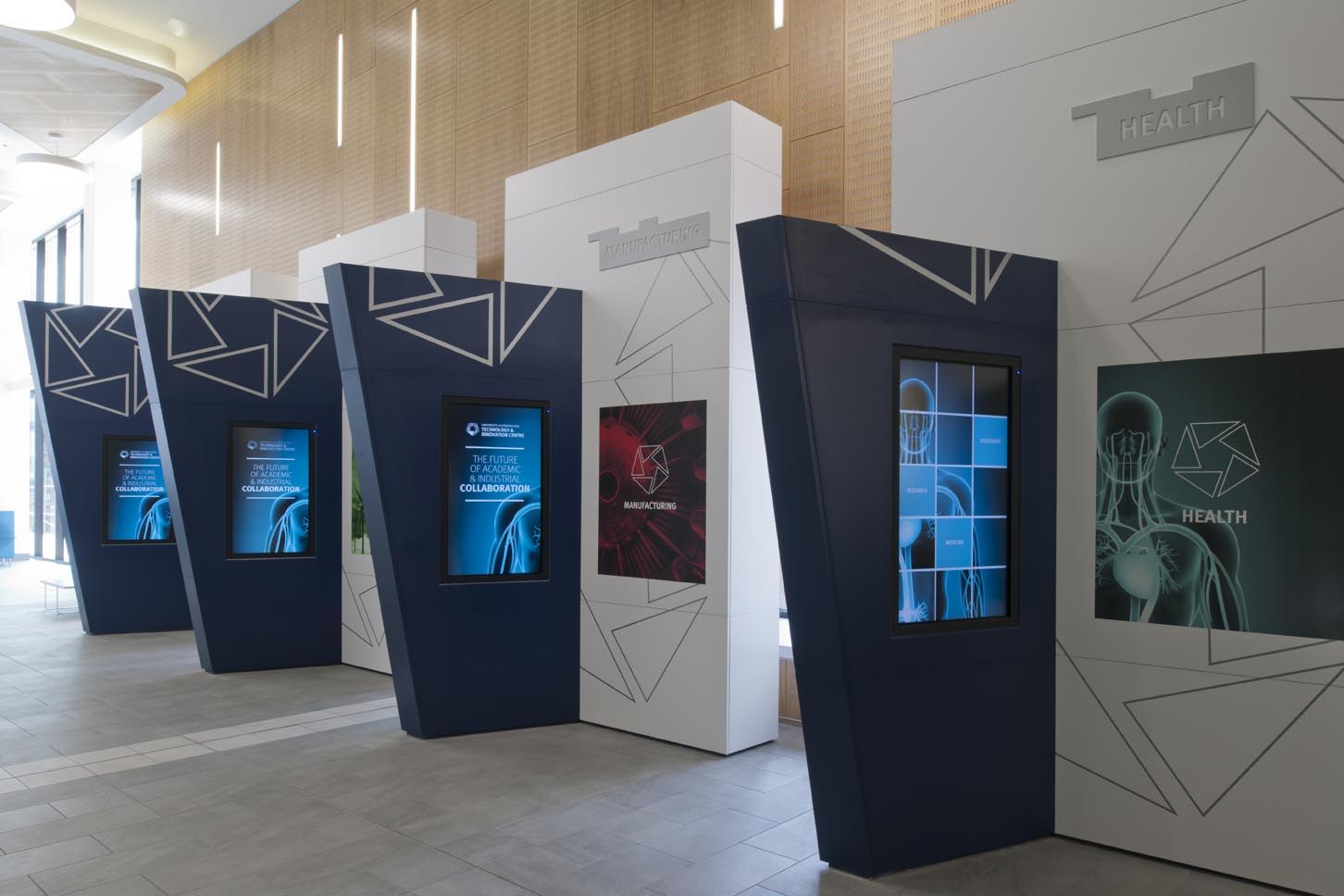 A row of modern digital kiosks with geometric designs and screens displaying graphics related to health and science in a bright, contemporary indoor space.