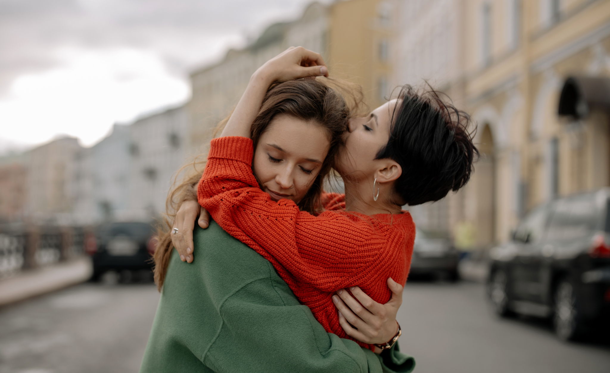 Two women hug while in the street. One is kissing the other on the head while they close their eyes.