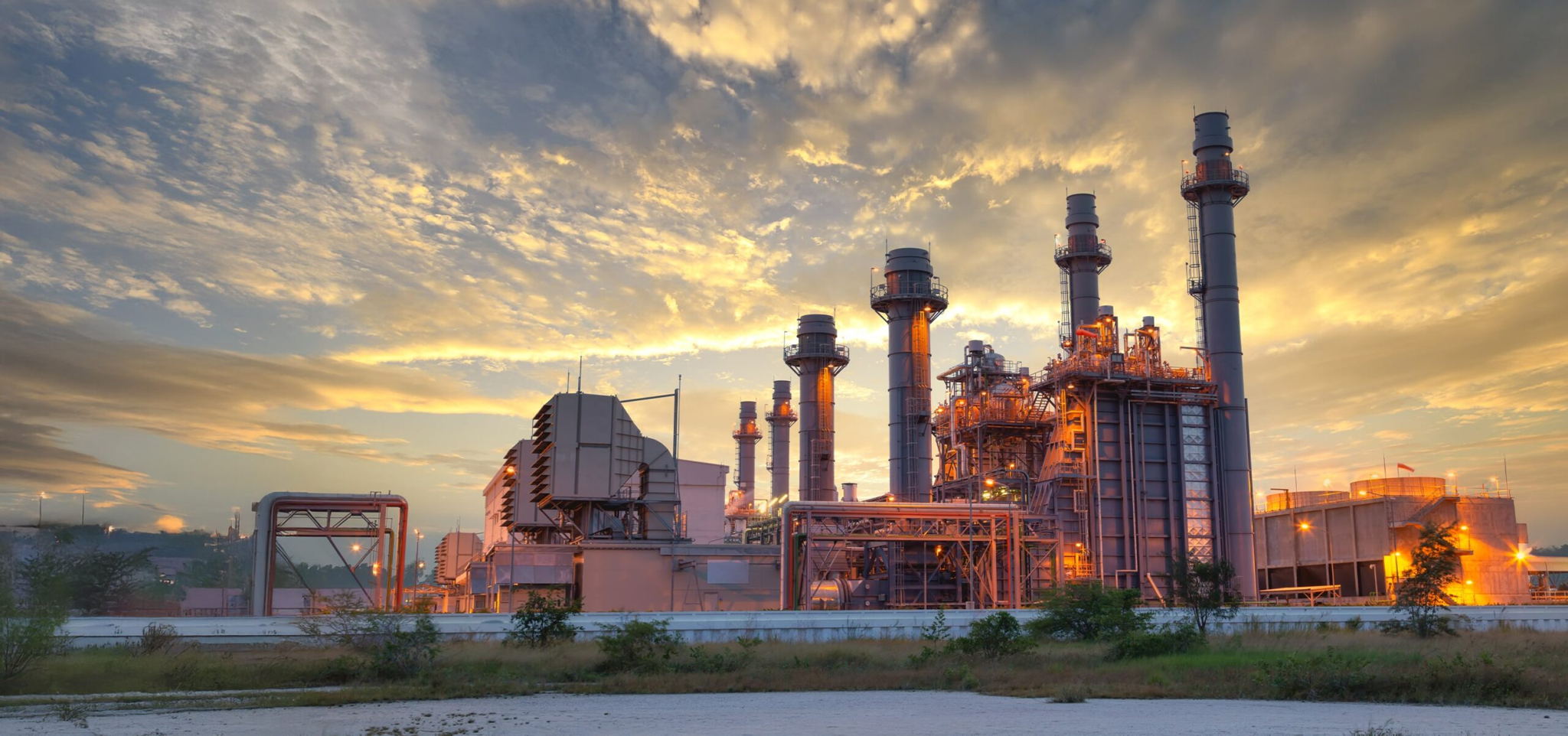 A large industrial power plant with multiple tall smokestacks and metal structures is set against a dramatic sunset sky, with light clouds and vegetation in the foreground.