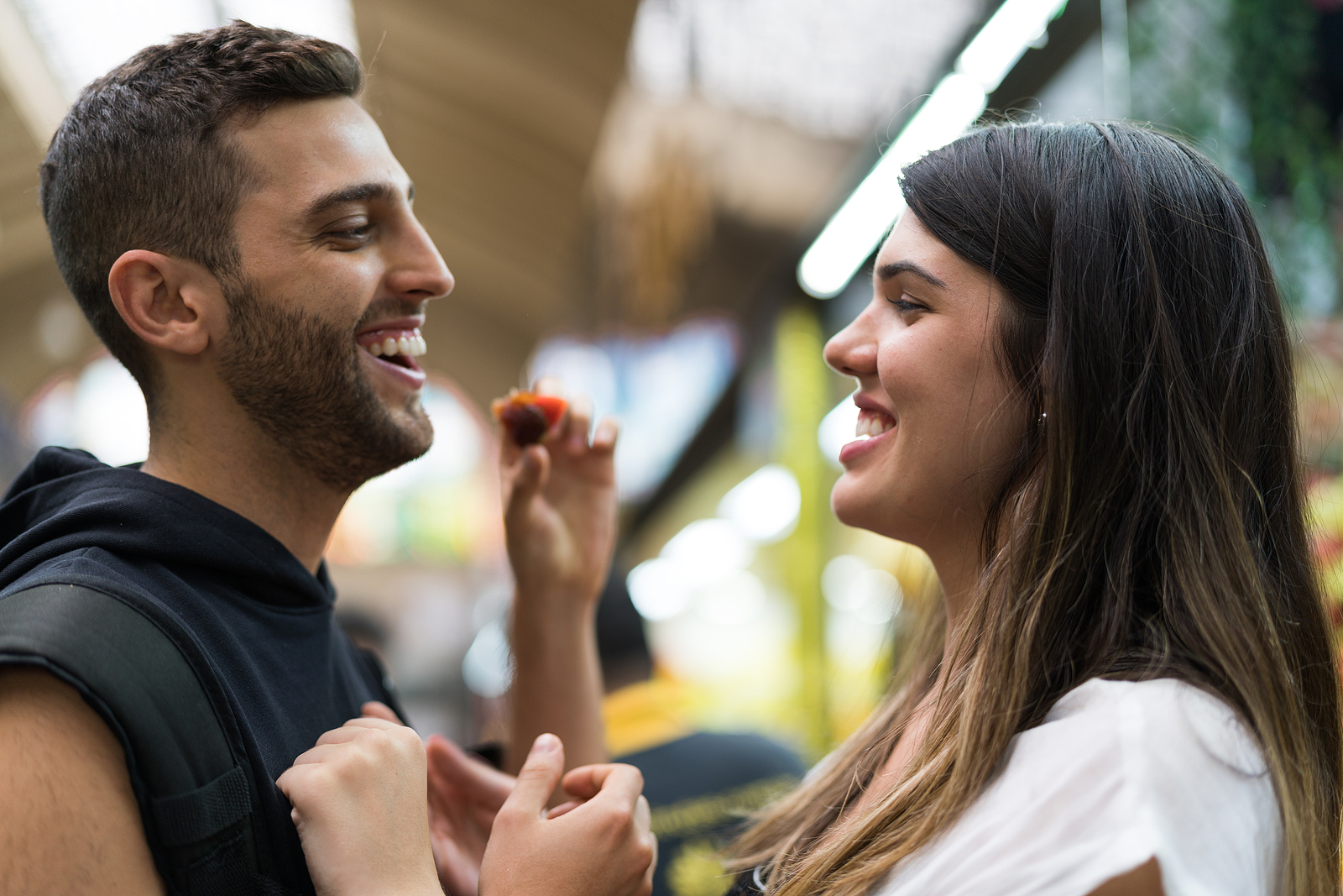An attractive man and a woman share an intimate moment in public. Both laugh while she gives him a bite of a snack.