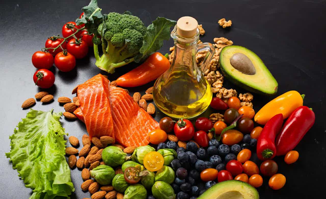 Assorted healthy foods including salmon, nuts, olive oil, and fresh vegetables on a dark surface.