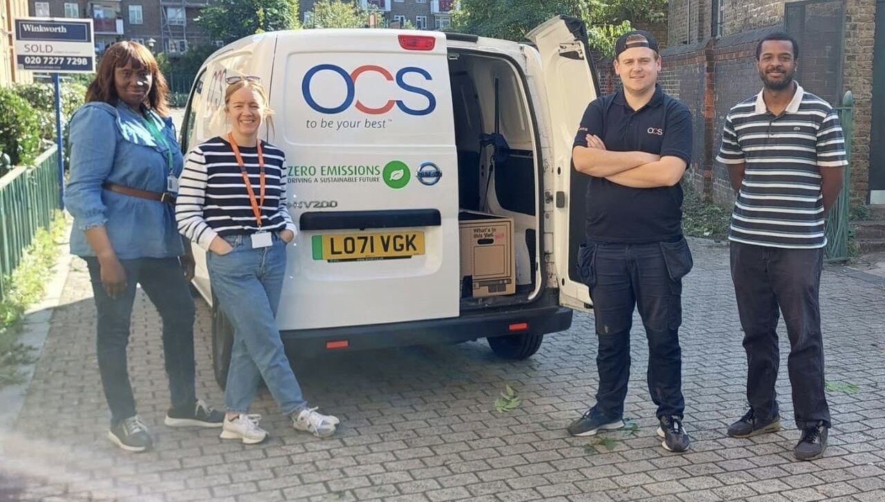 Four people stand smiling beside an OCS-branded white van with open back doors on a cobblestone street, with buildings and greenery in the background. The van displays “Zero Emissions” and a UK license plate.