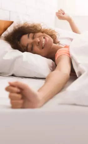 woman with curly brown hair, wearing an orange shirt, waking up and stretching in her bed