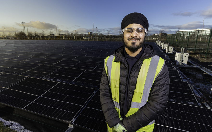 A man wearing a turban, glasses, and a yellow safety vest stands smiling in front of a large field of solar panels under a blue sky.