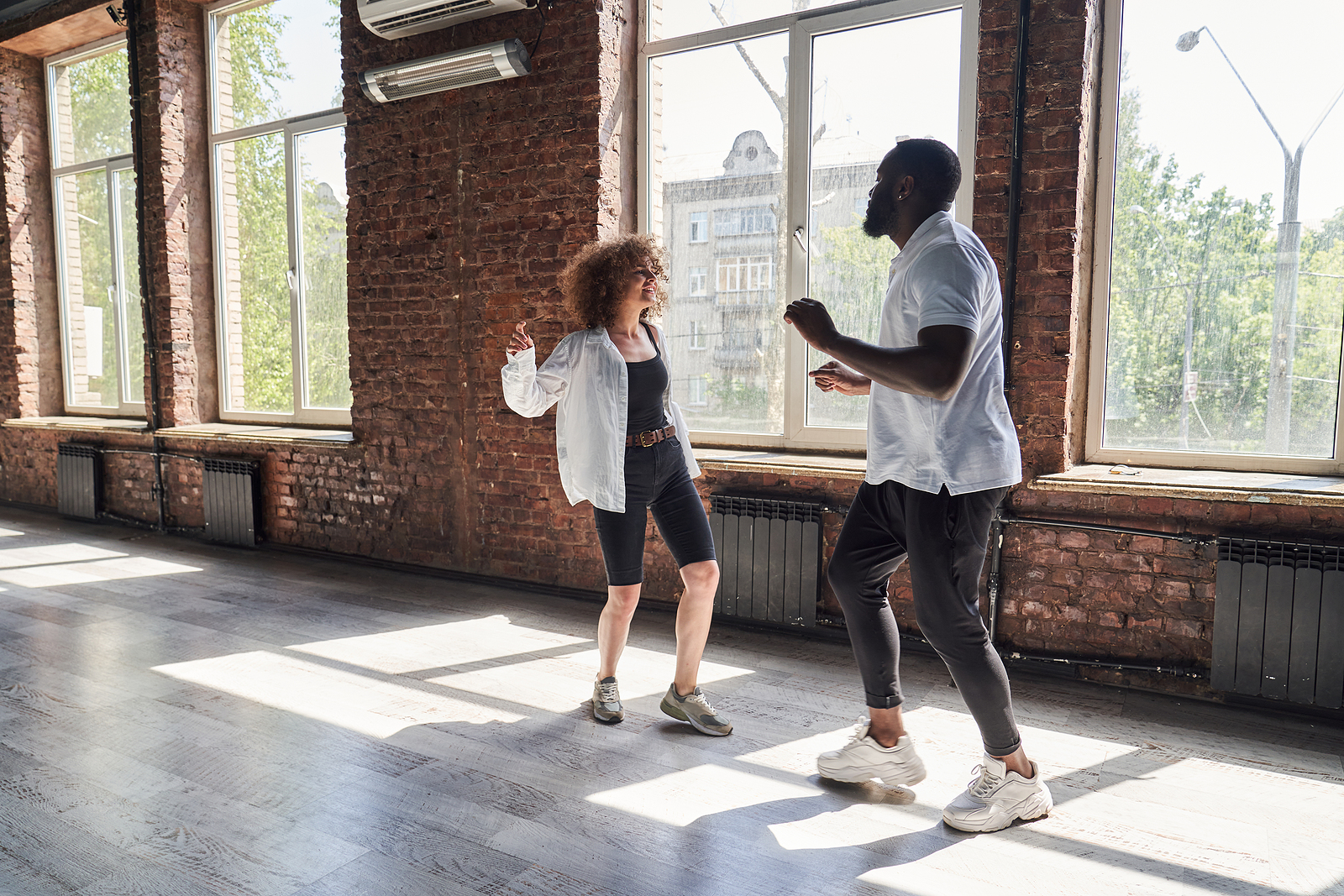 Photo of young hip black couple inside a brick room with open windows dancing together smiling.