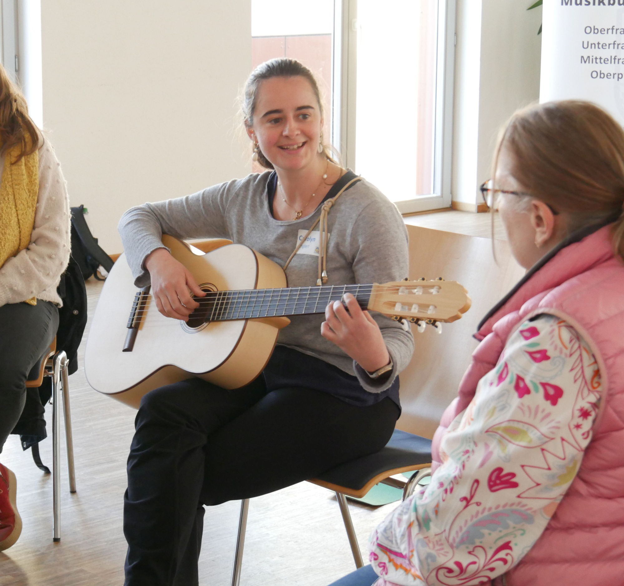 Eine Frau sitzt auf einem Stuhl und spielt Gitarre, eine andere Frau sieht ihr dabei zu. 