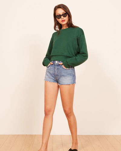 Front of woman wearing green organic cotton sweatshirt, light indigo cropped shorts and black sunglasses all from sustainable fashion brand Reformation