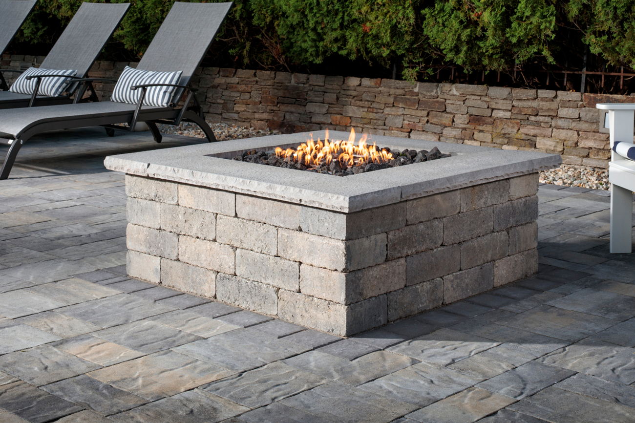 Weston Square Fire Pit on a patio made with vantage pavers, with patio chairs and a retaining wall in the background.
