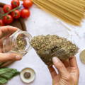 A glass spice jar being refilled from a plastic pouch filled with a leafy green spice