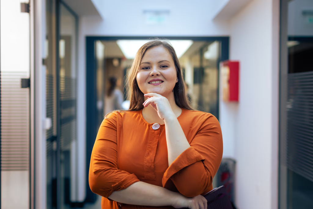 A woman in an orange shirt stands confidently in a modern hallway, smiling with her hand resting on her chin. Glass doors and blurred office elements are visible in the background.