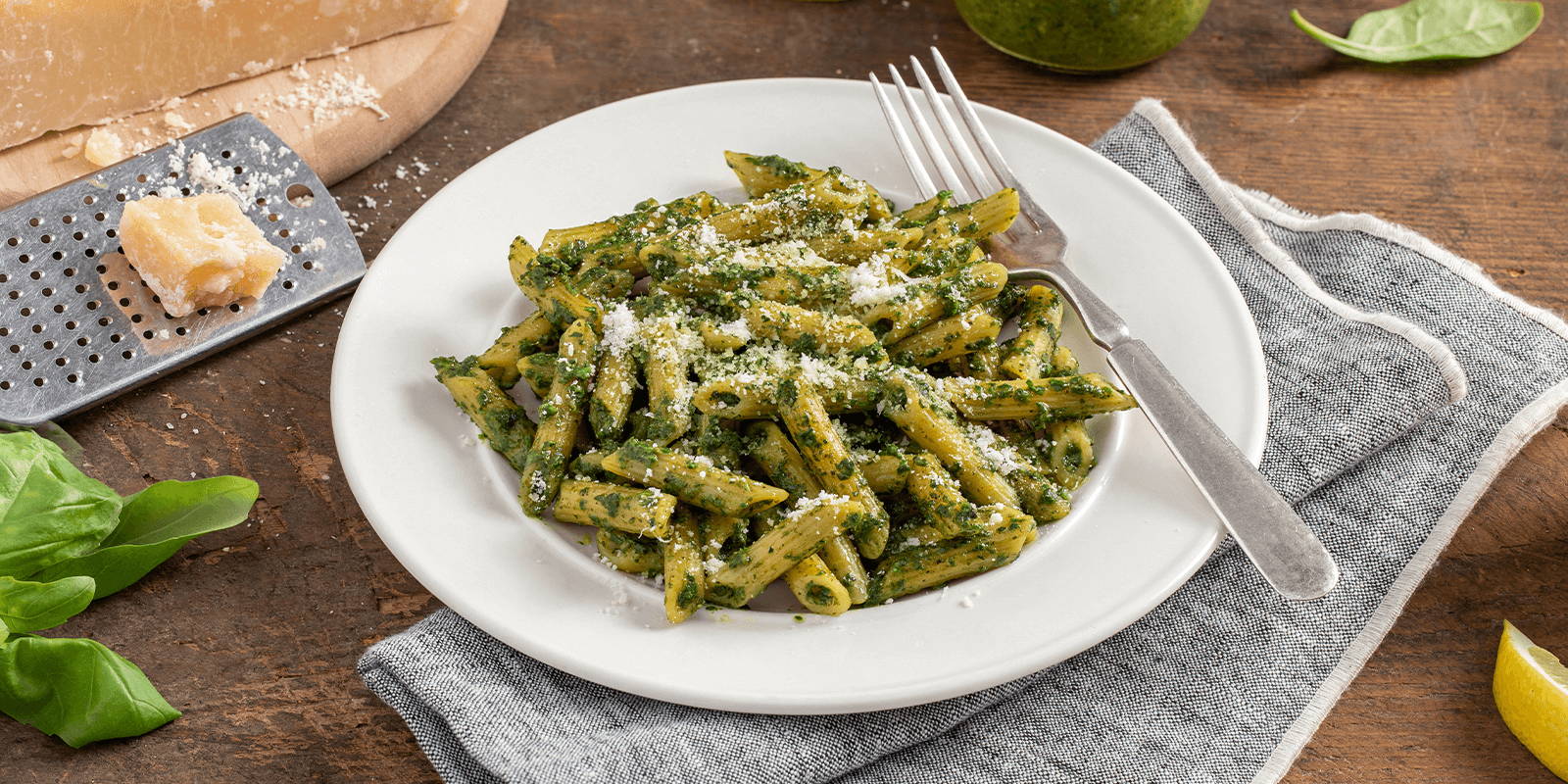 A plateful of Spinach-Basil Pesto With ZENB Penne Pasta atop a wooden table, styled with parmesan, basil leaves, and lemon slices.