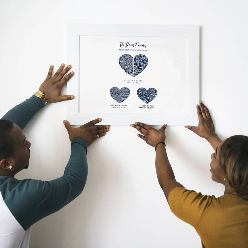 Bride and groom smiling at each other, holding a framed print with overlapping red and gray fingerprint hearts. Text below reads "Hillary + Brian" and wedding details.
