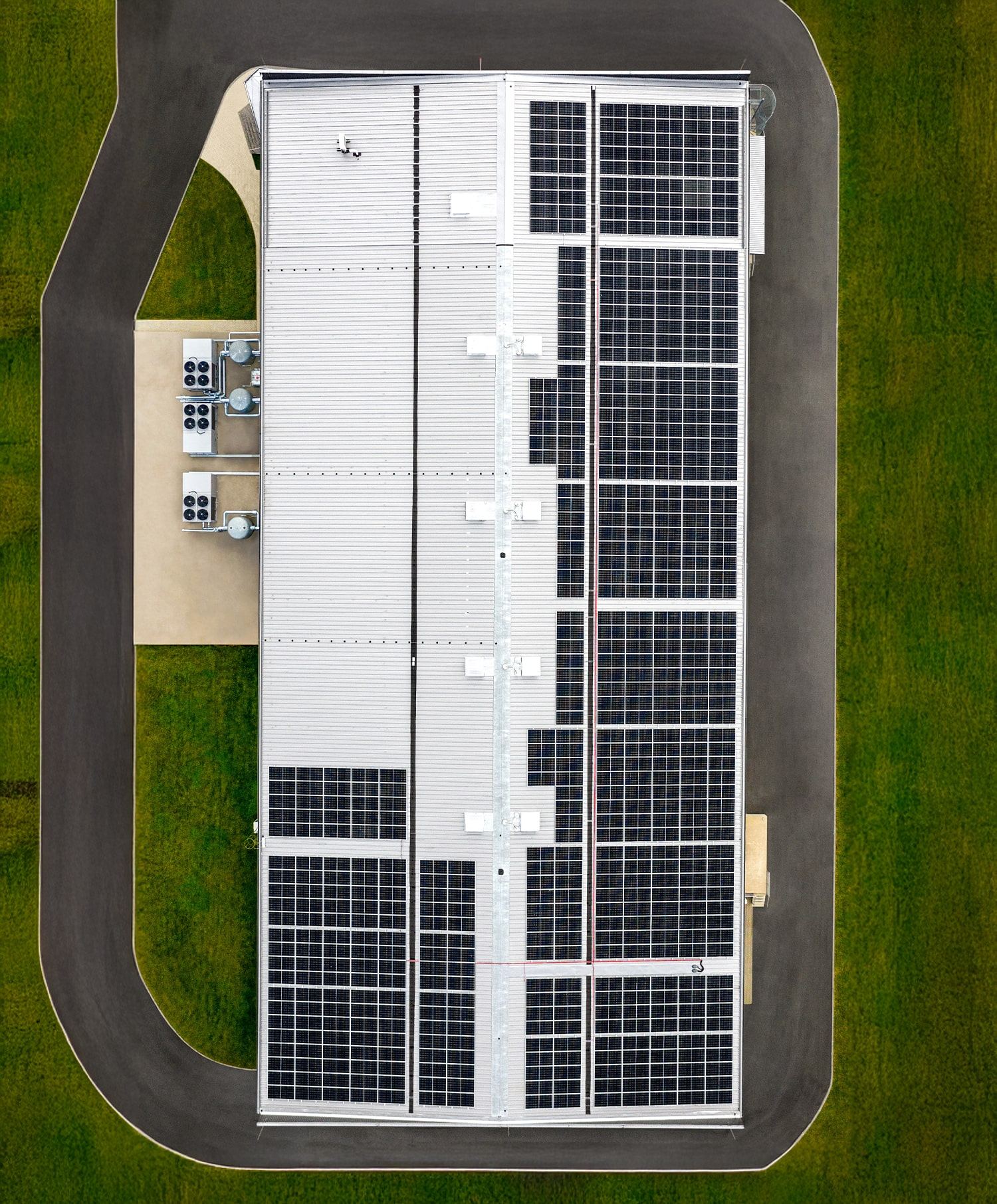 Aerial view of a large rectangular building with a roof covered in solar panels, surrounded by a paved road and green grass. HVAC units are visible on a platform next to the building.