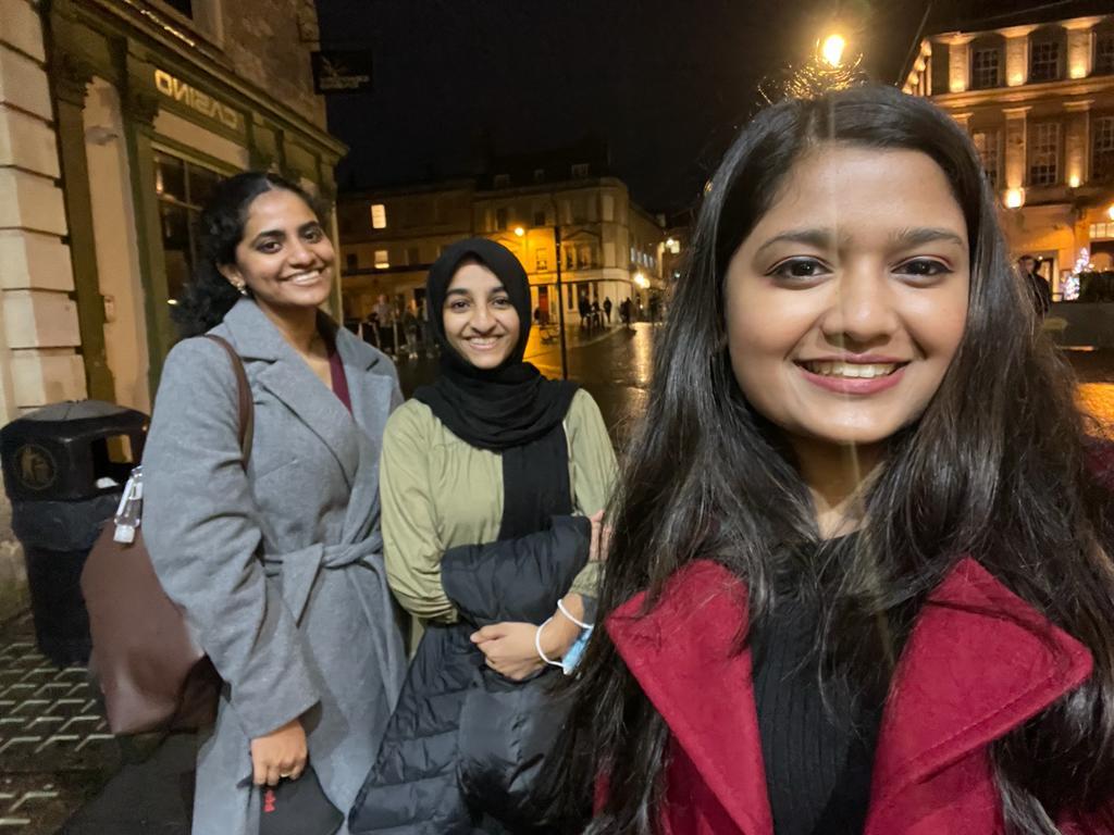 Gayathri smiling with her friends lit by a streetlight. They are wearing winter clothing.