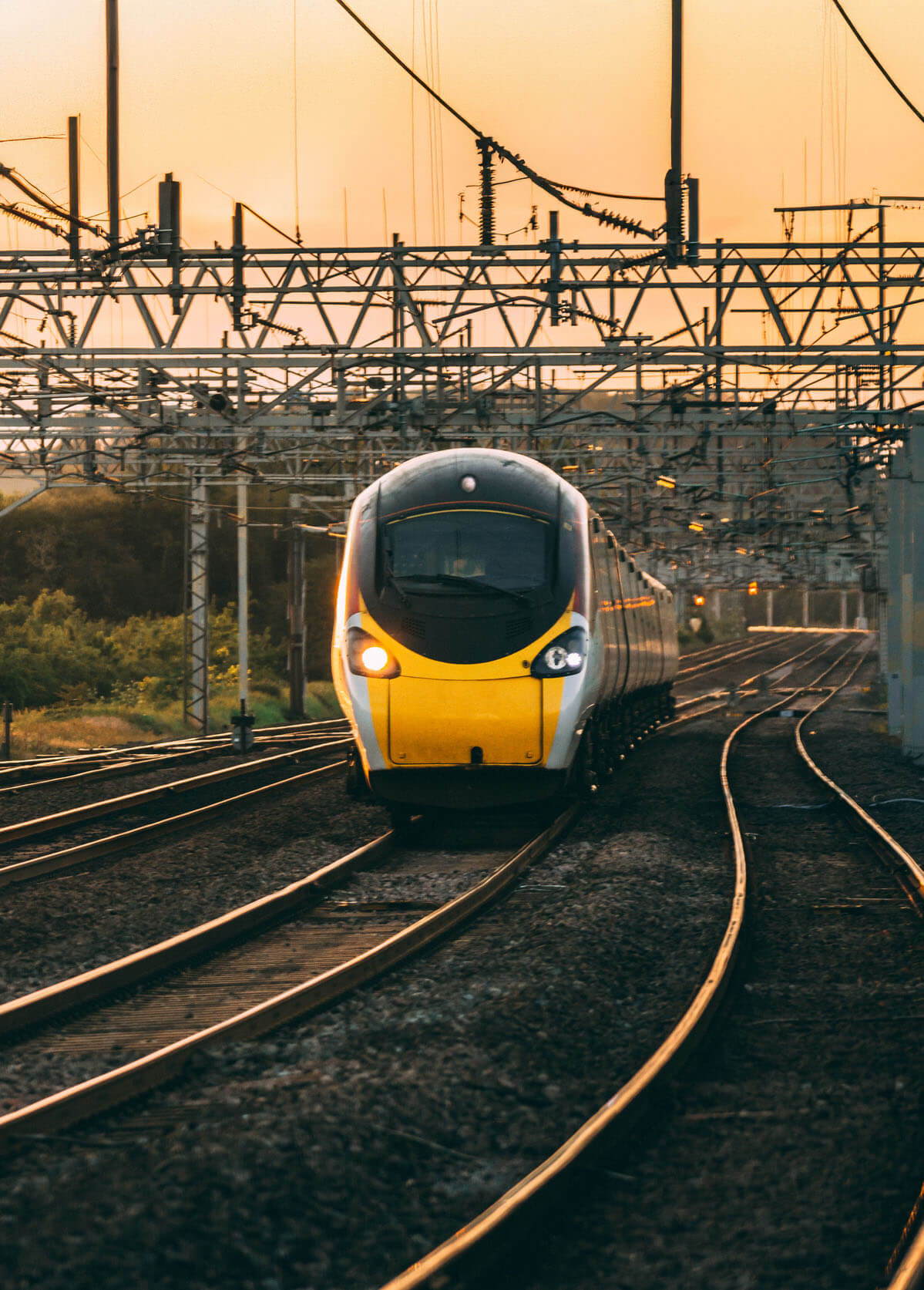 A sleek passenger train with a yellow and black nose travels down railway tracks at sunset. Power lines crisscross overhead, and the warm glow of the sun illuminates the scene, casting long shadows on the tracks.
