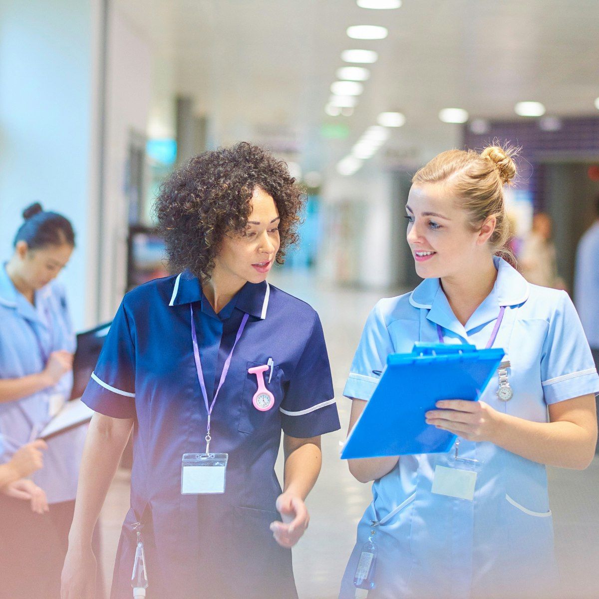 Two nurses in uniforms walk and talk in a brightly lit hospital corridor. One holds a blue clipboard, and both wear ID badges. Other healthcare staff work in the background.