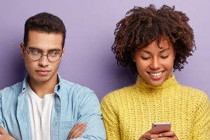 A man with his arms crossed looks over a his friend who is smiling looking down at her phone. He has an annoyed expression.