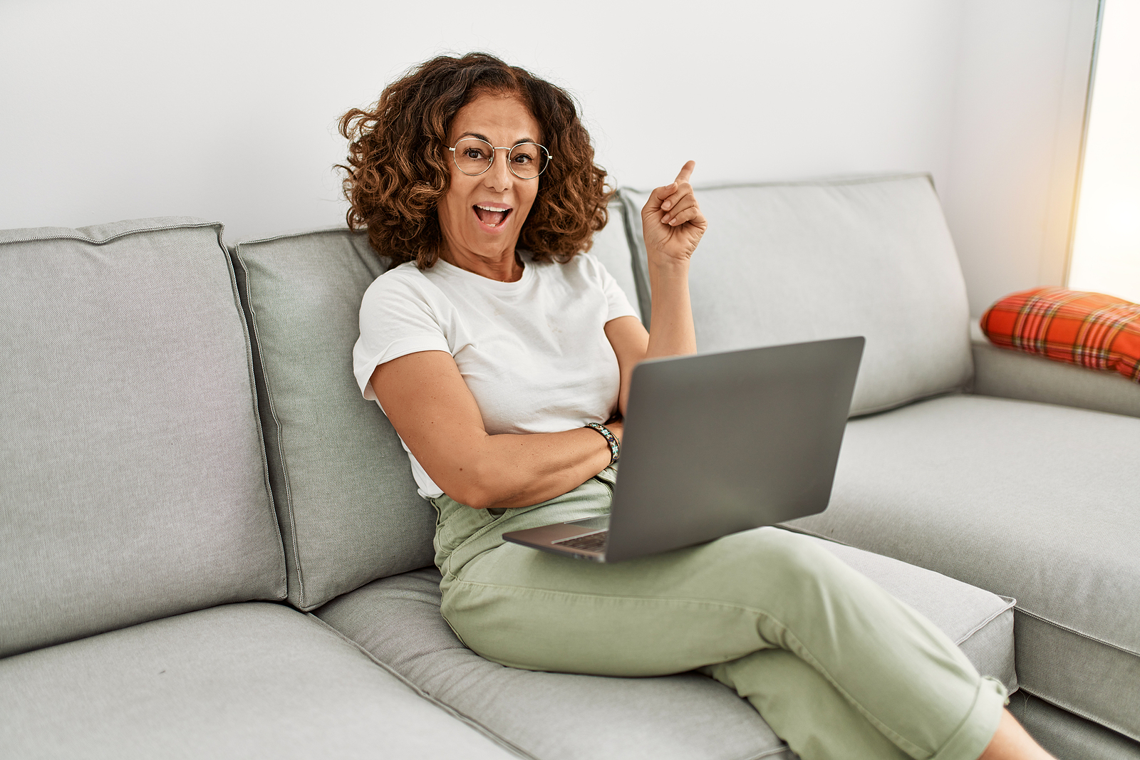 A middle aged latin woman smiles while working on her laptop on her couch with a finger pointing outwards.