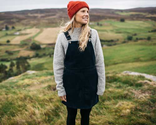 Woman wearing Finisterre Black cord dungaree dress on top of grey knit and an orange beanie hat in a countryside landscape