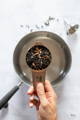 A hand holds a half-cup measuring cup filled with dried elderberries and spices over a metal saucepan on a white surface.