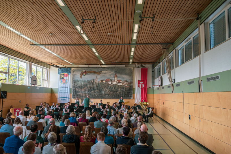 In einer Turnhalle musiziert ein Blasorchester in Tracht vor Publikum