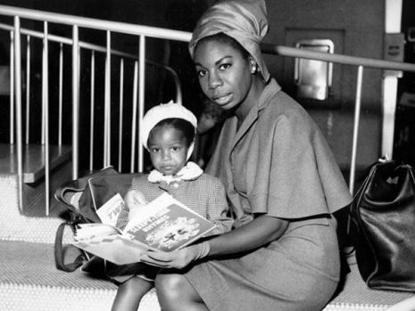 Nina simone with her daughter, sitting together and reading a book. Both are wearing dresses.