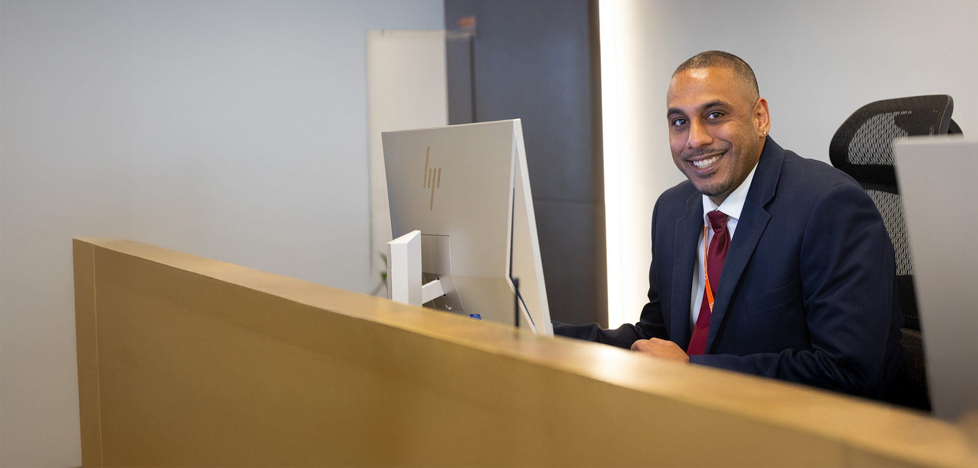 A man in a dark suit sits at a modern reception desk, smiling at the camera, with a computer monitor in front of him and an office background behind.