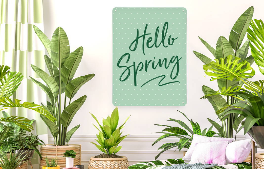 A magnetic notice board wall art panel with a green polka dot design on a wall in an interior with lots of plants and greenery. Hello Spring is written on the board in a green script.