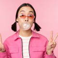 Model standing in fron of a pink background, holds up both hands with fingers in the victory sign. Model has pink glasses, hair in bunches, pink jacket and is blowing a bubble gum bubble.