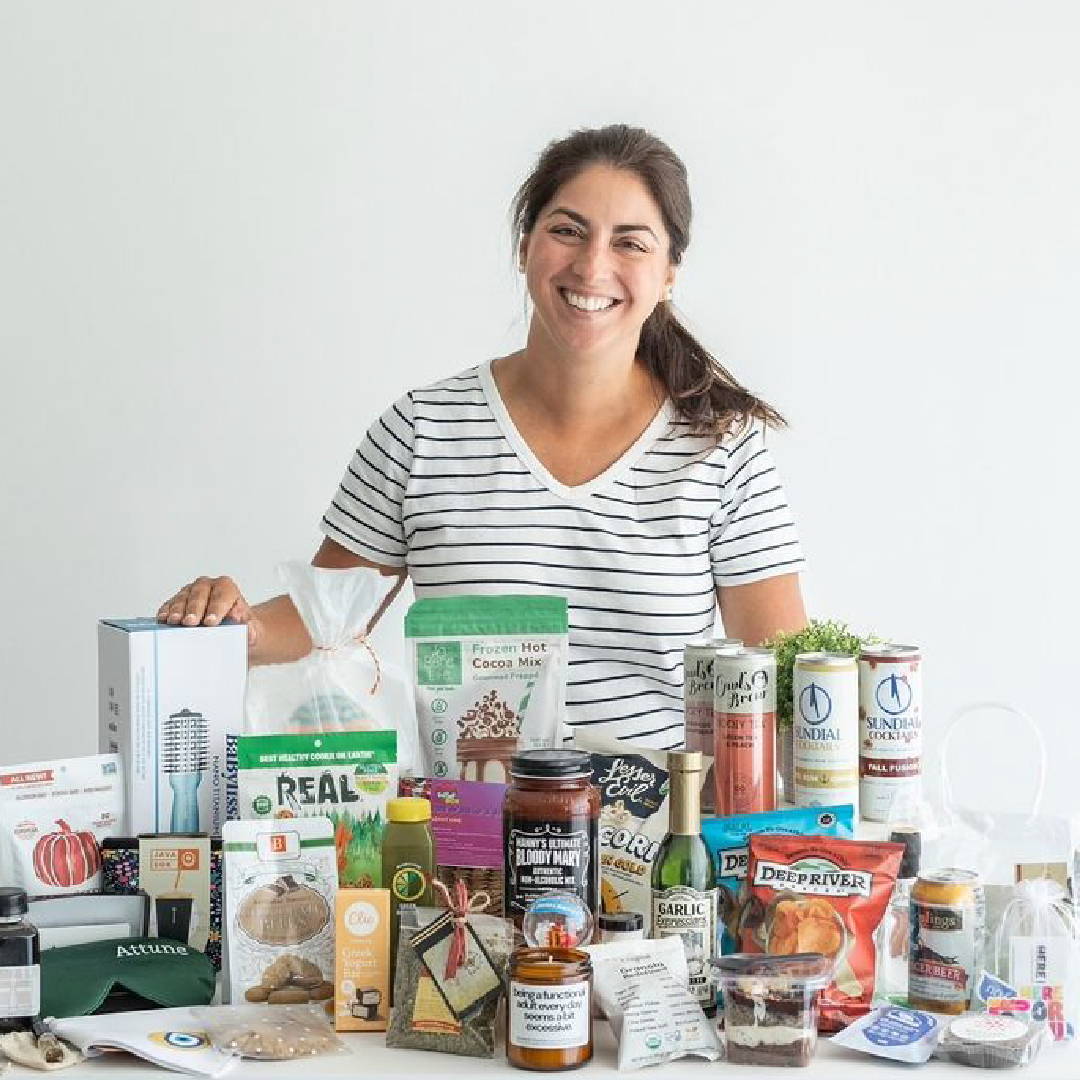 A smiling woman stands behind a table filled with various organic, plant-based products, including a bag of Bare Life Frozen Hot Cocoa Mix. The assortment showcases healthy, gluten-free, dairy-free, and vegan items, reflecting the blog's focus on allergen-friendly, simple, and sustainably sourced ingredients.