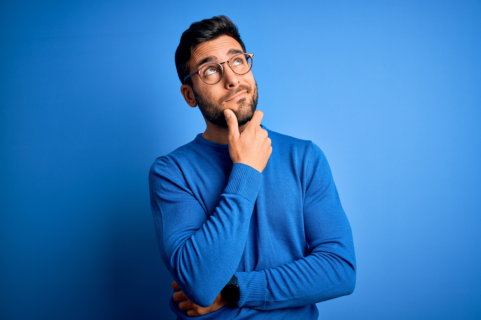 An attractive guy wearing glasses smiles and holds his hand on his chin and looks deep in thought.