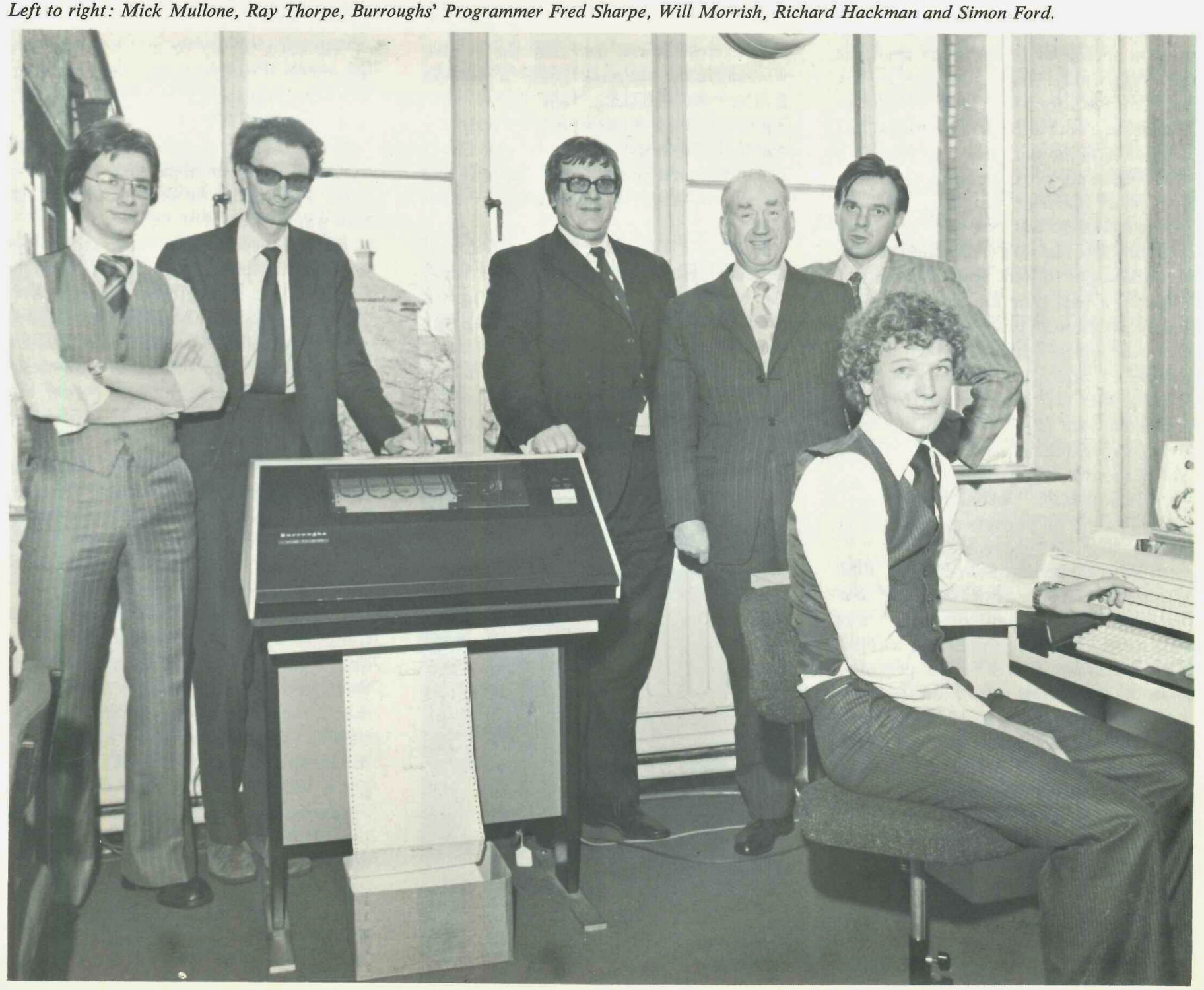 Six men in formal attire pose in an office with large windows. One is seated at a desk with a computer, while the others stand behind a vintage computer terminal. The setting appears to be from the late 20th century.