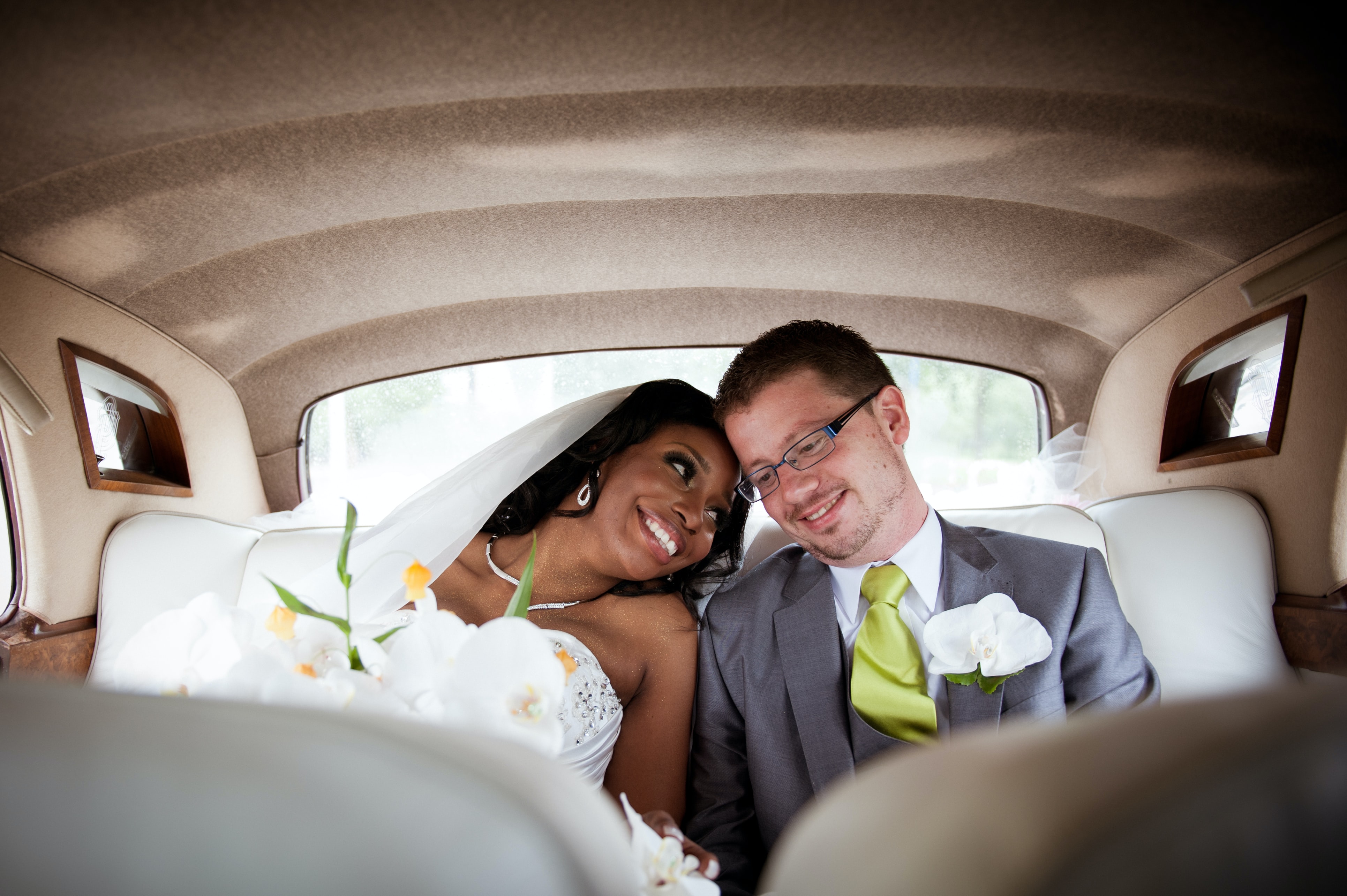 A multi racial couple sits in the back of a car smiling to eachother in their wedding outfits.