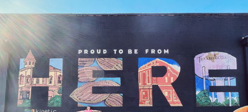 Vibrant mural with "HERE" and "TEXARKANA," showcasing local pride, joy, and creativity; person celebrates community spirit.