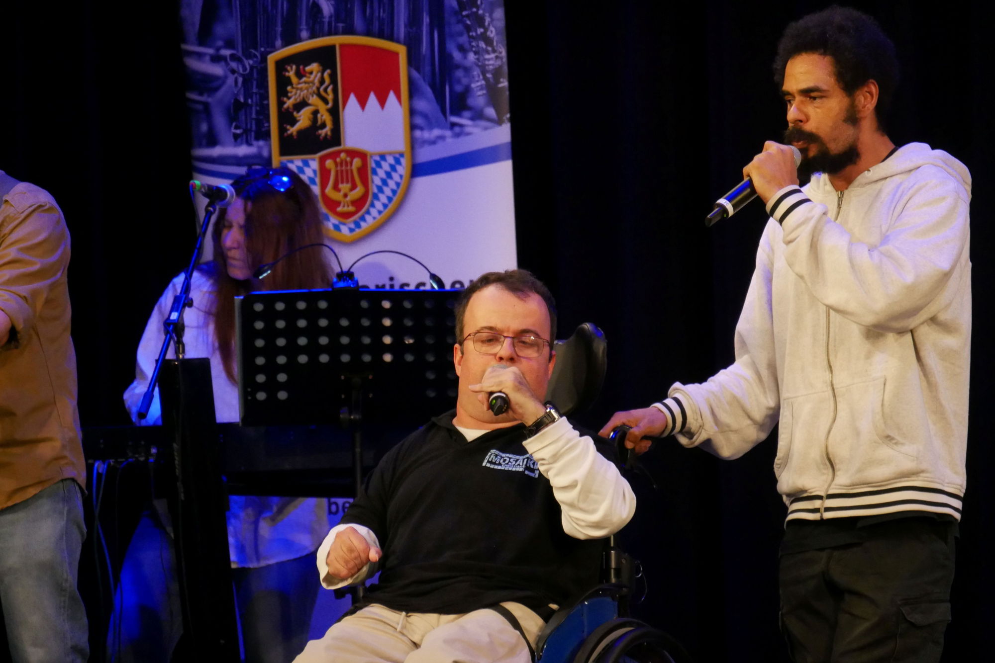 Zwei Männer singen gemeinsam auf einer Bühne. Einer der beiden Männer sitzt im Rollstuhl. 