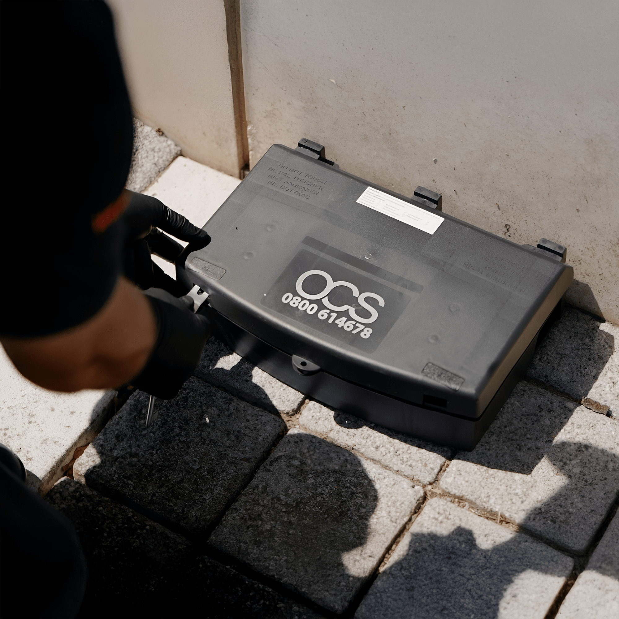 A person wearing black gloves is handling a black plastic box labeled OCS 0800 614678, placed on outdoor paving stones next to a wall.