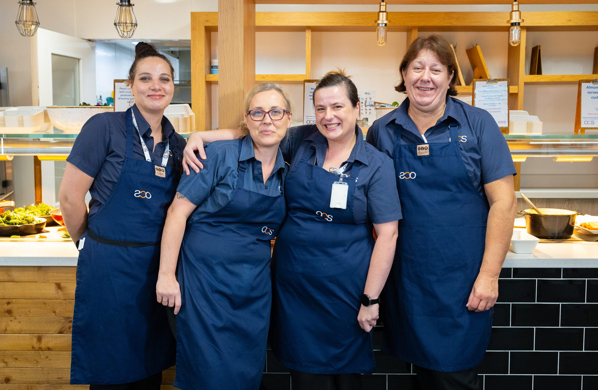 Four smiling women wearing matching blue uniforms and aprons stand together in front of a cafeteria counter, posing for the photo. There are plates of food and serving dishes visible in the background.