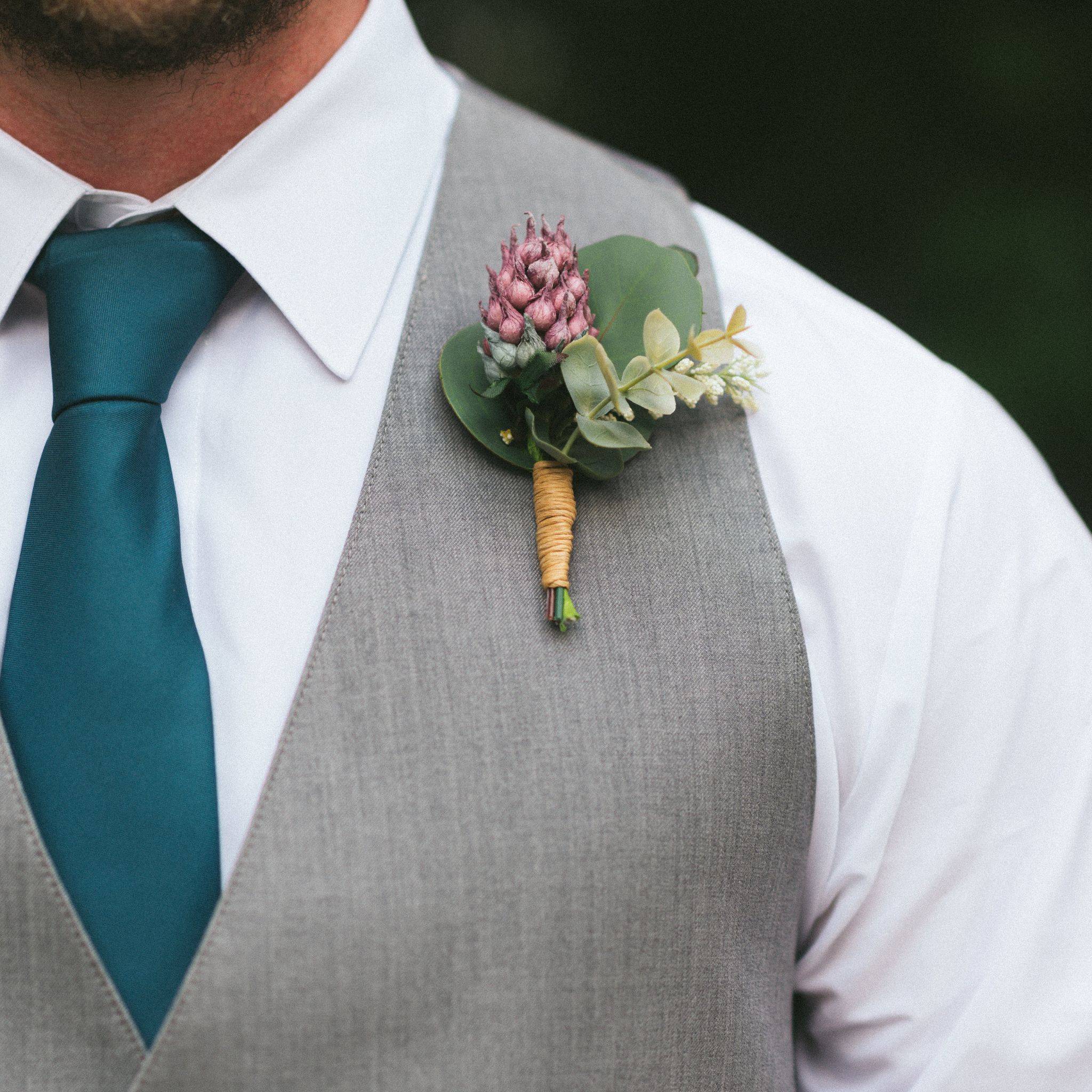 A boutonniere made of purple thistle, baby eucalyptus and silver coin eucalyptus is pinned to a groom's chest who is wearing a gray vest on a white shirt with a teal blue tie