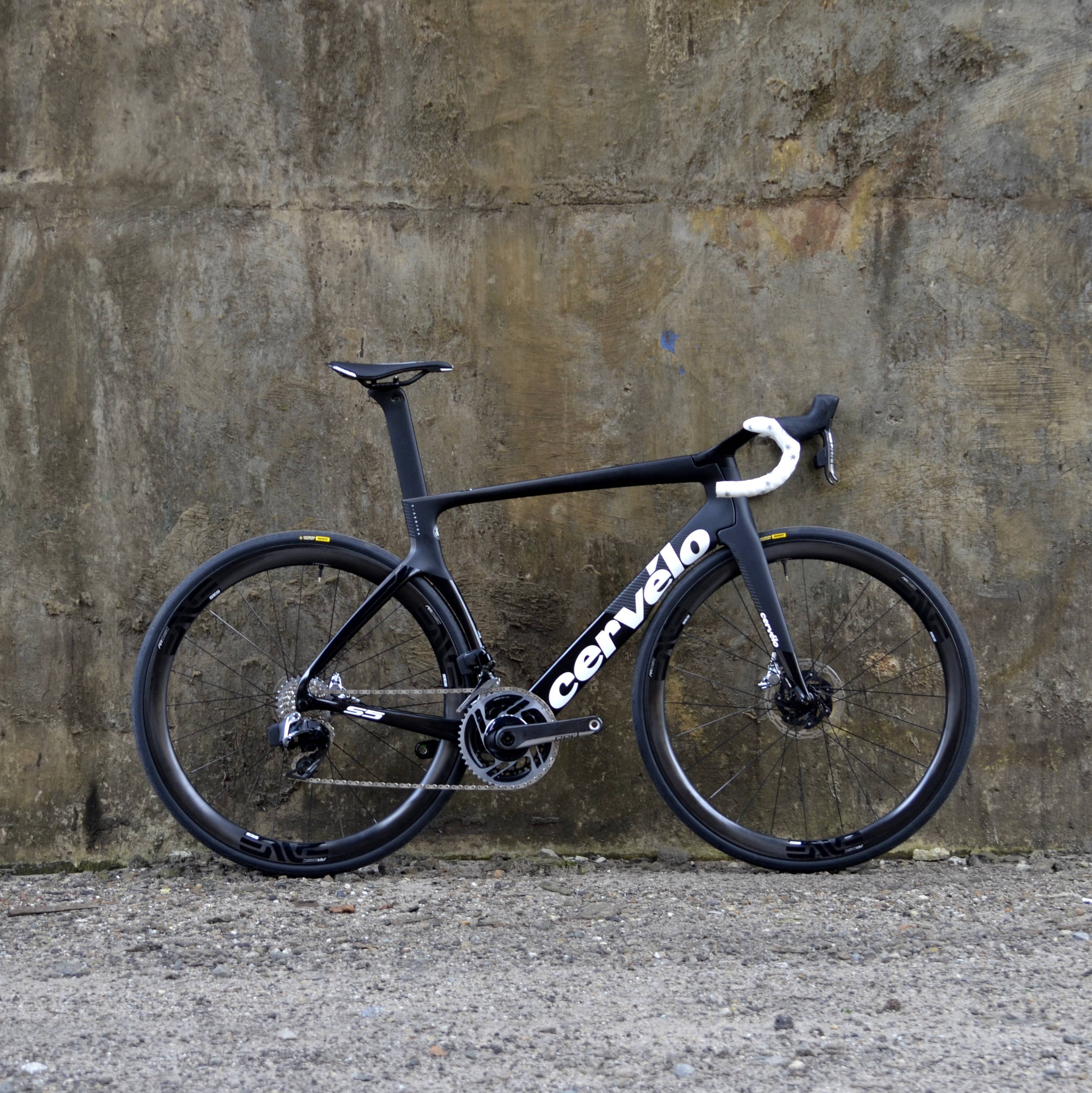 Cervélo S5 aero racefiets in zwart zijaanzicht tegen een verweerde betonnen muur, met diepe carbon velgen, schijfremmen en wit stuurlint op een ruwe grindondergrond.