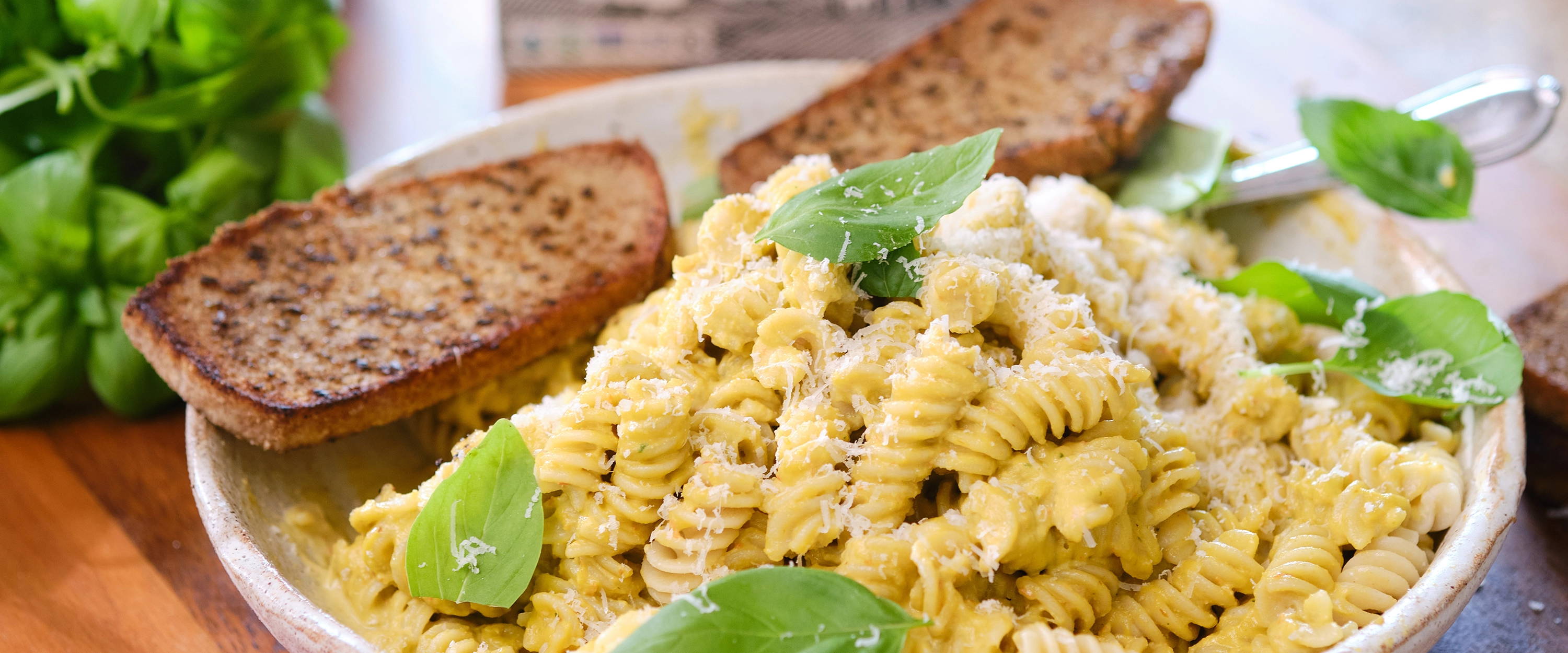 A bowl of fusilli pasta with tomato sauce, almonds, and basil, served with two slices of garlic bread on the side, surrounded by other breakfast items on a wooden table.