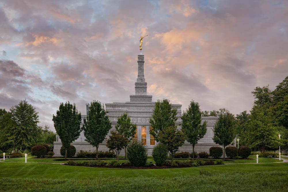 LDS art photo of the Palmyra New York Temple surrounded by green grounds and trees during sunrise.