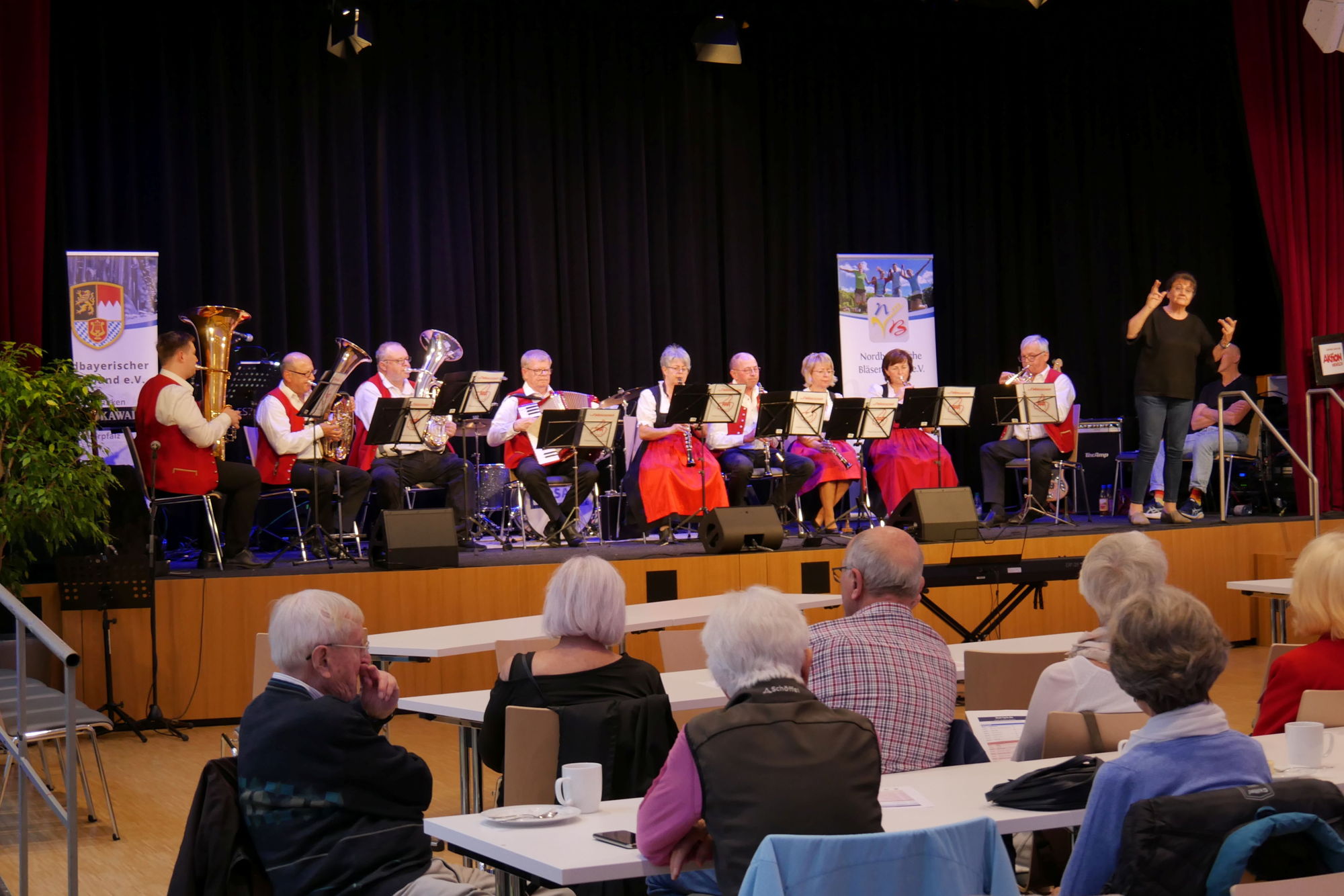 Eine Kapelle in Tracht musiziert auf einer Bühne vor Publikum. Daneben steht eine Gebärdendolmetscherin und übersetzt die Musik. 