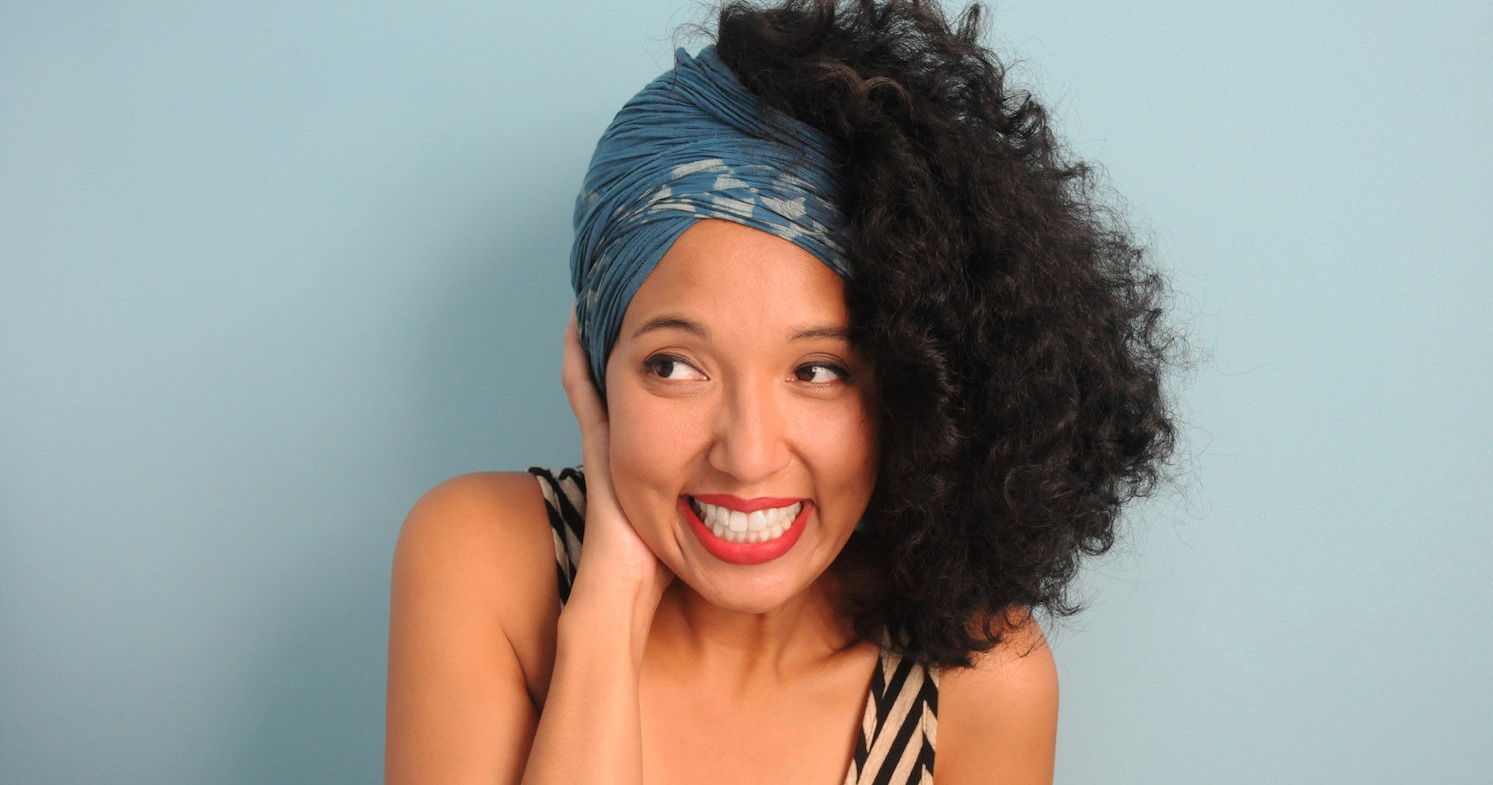 A young multi ethnic woman wearing a head wrap and curl hair, smiles big while holding her face and looking to the side nervously.
