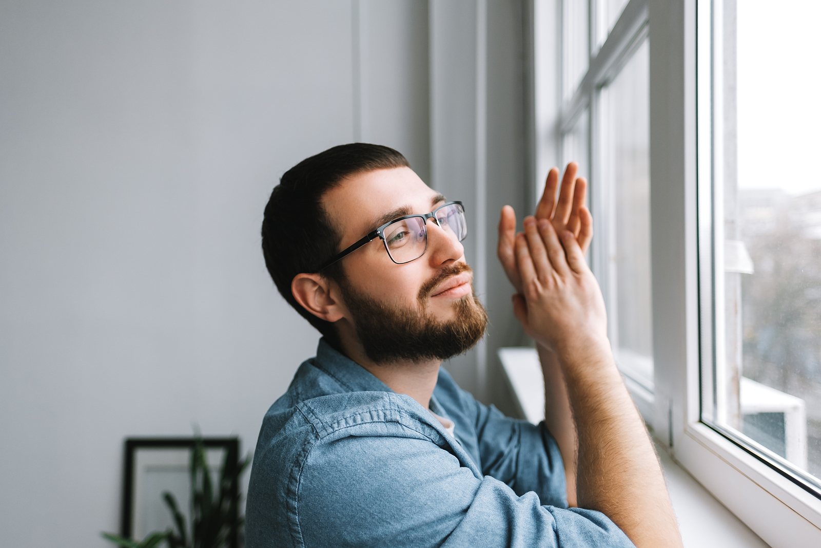 White man with a beard and glasses, day dreaming near a window looking out.