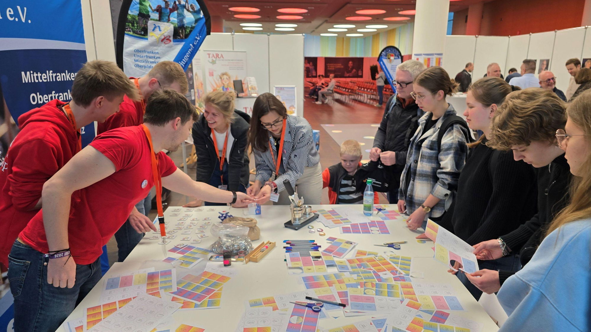 Einige junge Erwachsene und Kinder stehen um einen Tisch mit vielen bunten Ausdrucken und basteln Buttons. 