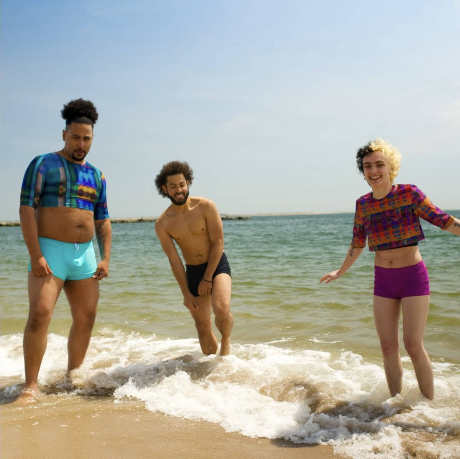 Three  nonbinary models wear Play Out Apparel Swim Trunks, Swim Shorts, and cropped swim rash guards while smiling and standing in the ocean.