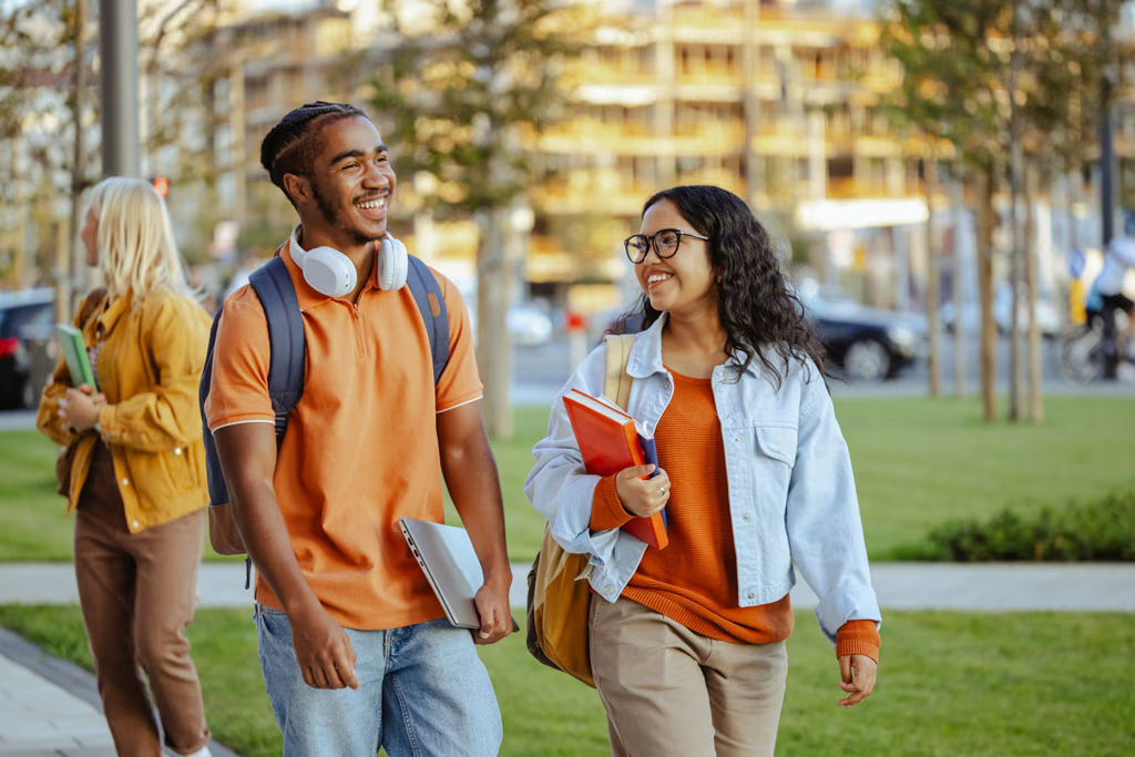 Two college students walk and smile while carrying books and laptops on a grassy campus. A person walks in the background. The scene is bright and outdoors, with buildings and trees visible behind them.