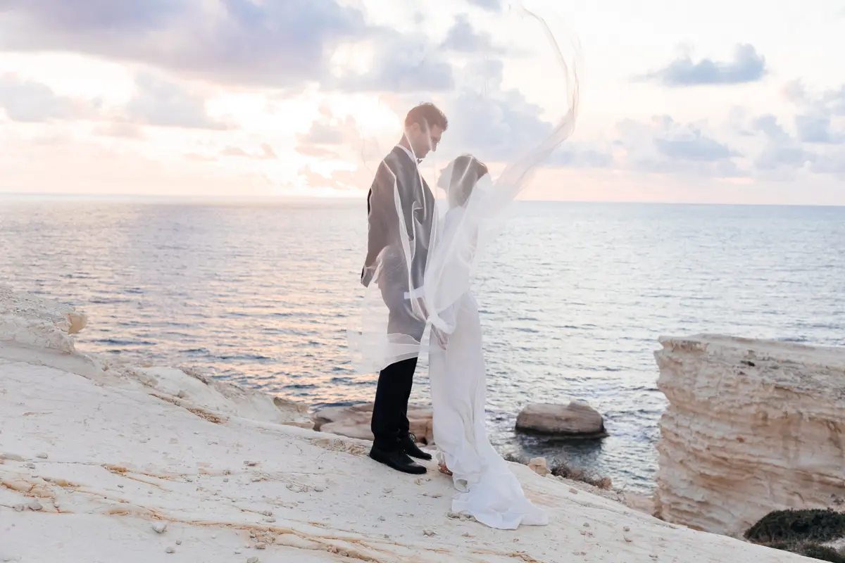 Un couple se tient sur une falaise isolée, dominant la mer Méditerranée, le voile de la mariée flottant élégamment dans la brise marine alors qu'ils échangent un sourire complice.